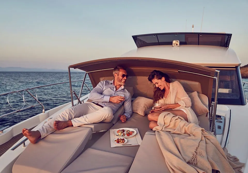 A man and woman relax on the deck of a yacht, smiling and enjoying drinks. Plates of food are on a table beside them. The sea and horizon are visible in the background under a clear sky.
