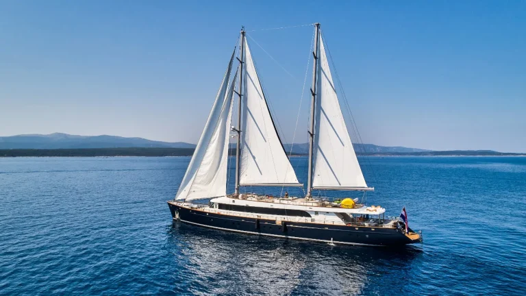 A luxurious two-masted sailing yacht with white sails glides on calm blue water, with a clear sky and distant mountains visible in the background.