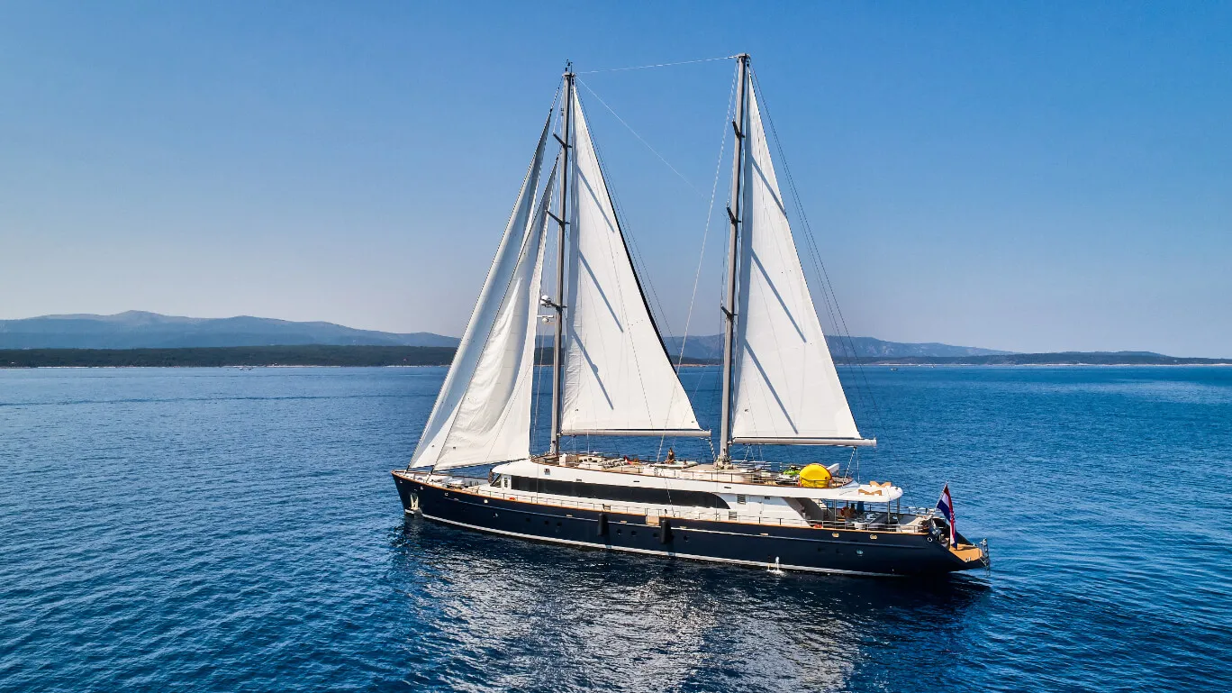 A luxurious two-masted sailing yacht with white sails glides on calm blue water, with a clear sky and distant mountains visible in the background.