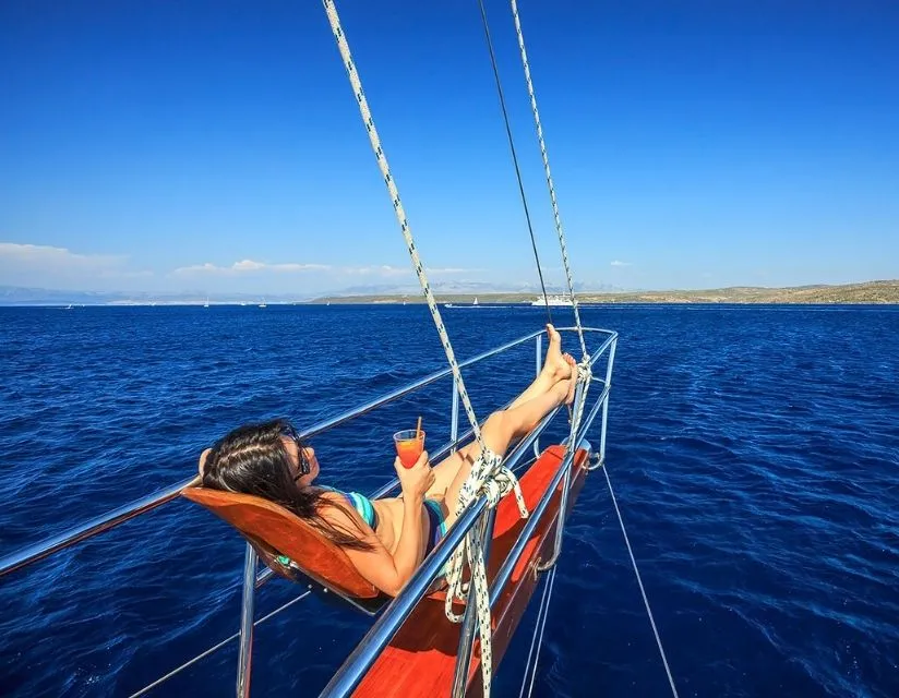 A woman relaxes in a hammock on the bow of a sailboat, holding a drink and looking out over deep blue water under a clear sky, with distant land visible on the horizon.