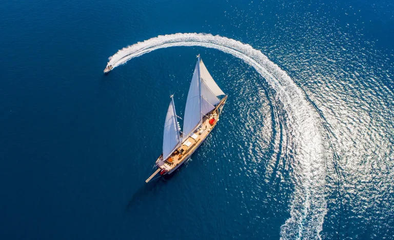Aerial view of a sailboat with white sails cruising on deep blue water, while a speedboat circles around it, creating a white arc of waves on the calm sea.