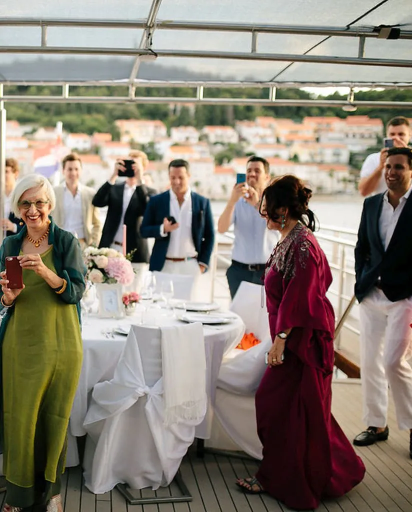 A group of people on a boat, dressed in formal attire, smile and take photos. Two women stand in the foreground, one in a green dress and another in a magenta dress, near a table set for a meal with white linens and flowers.