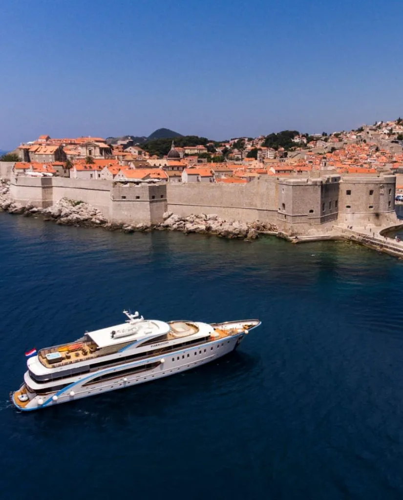 A white luxury yacht sails on deep blue water near the stone walls and orange rooftops of Dubrovnik’s historic Old Town in Croatia, under a clear blue sky.