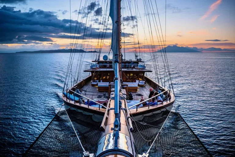 A view from the bow of a wooden sailboat at sunset, with calm blue water and distant islands on the horizon under a dramatic, colorful sky.