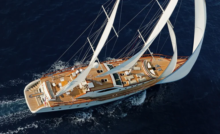 Aerial view of a large white sailboat with wooden decking, three tall sails, and outdoor seating areas sailing through deep blue water, creating small waves behind it.
