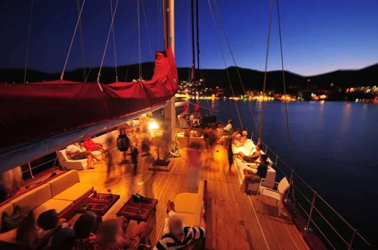 People relax and socialize on the deck of a sailboat at dusk, with warm lights illuminating the wooden deck and distant city lights reflecting on calm water in the background.