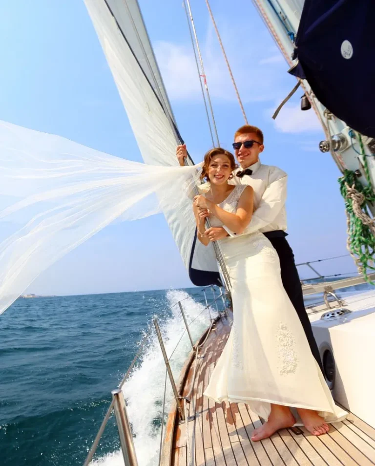A bride and groom smile and pose together on the deck of a sailboat at sea, with the bride’s veil blowing in the wind and waves splashing alongside the boat under a clear blue sky.