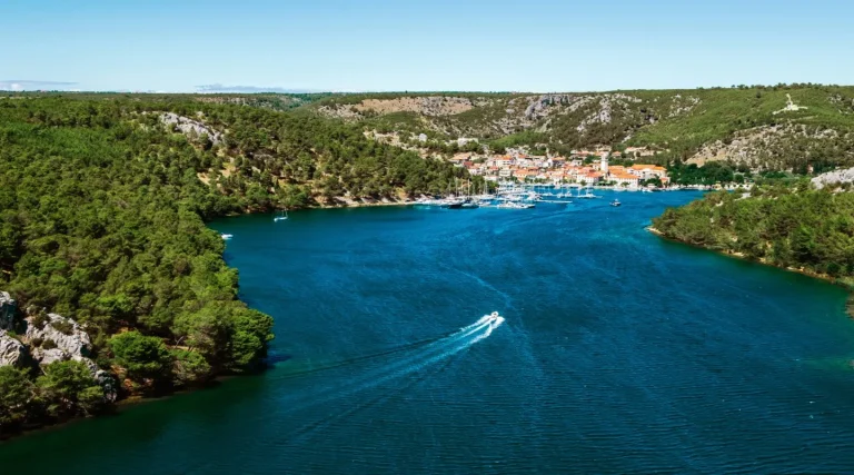 Aerial view of a green forested river valley with a small boat creating white wake on blue water, leading towards a coastal town with orange-roofed buildings nestled among hills under a clear sky.