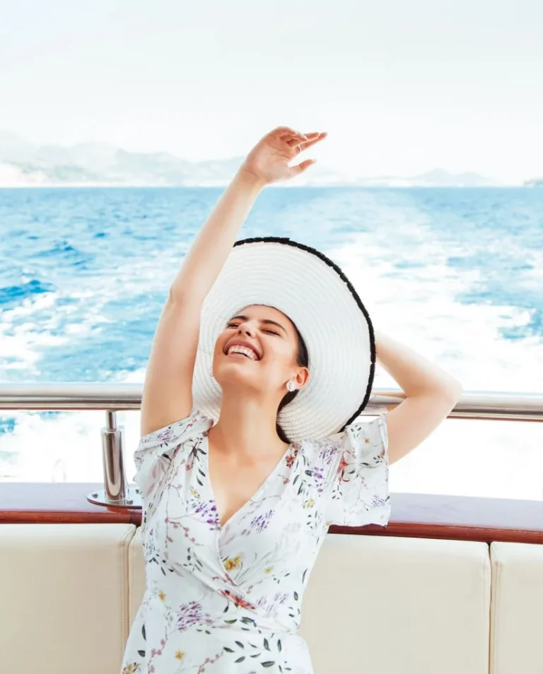 A woman in a floral dress and wide-brimmed hat sits on a boat, smiling and raising one arm joyfully, with the sea and distant hills in the background.