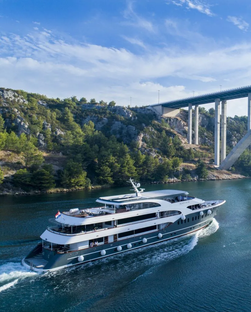 A modern white luxury yacht sails on a calm river beneath a tall bridge, surrounded by rocky hills and green trees under a partly cloudy blue sky.