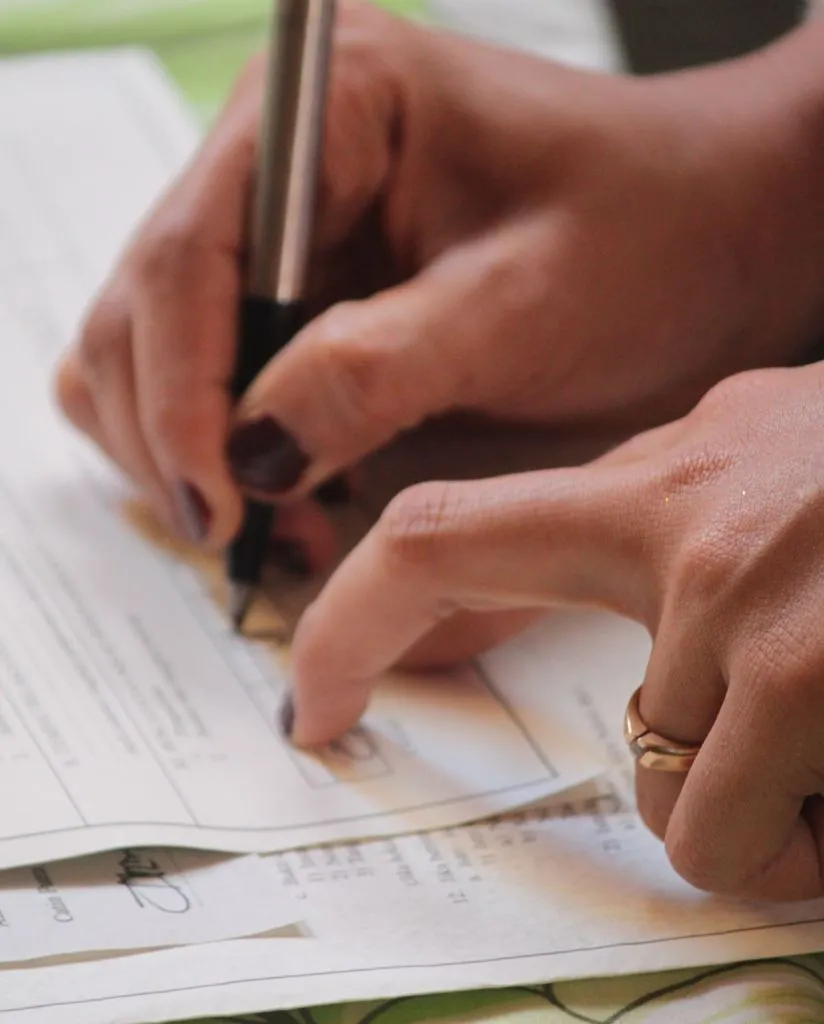 Close-up of a person’s hands filling out a form with a pen, wearing a gold ring on their left hand, with several sheets of paper visible underneath.