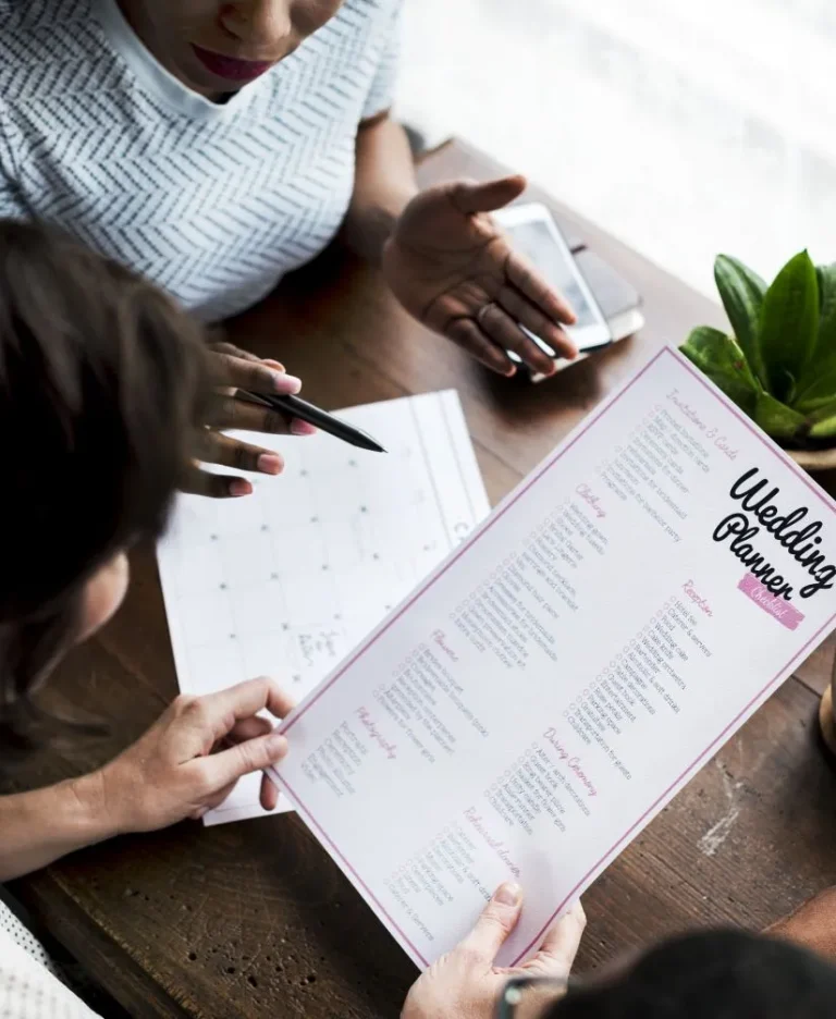 Two people sit at a table planning a wedding, one holding a Wedding Planner checklist and another pointing at a calendar, with a potted plant and smartphone on the table.
