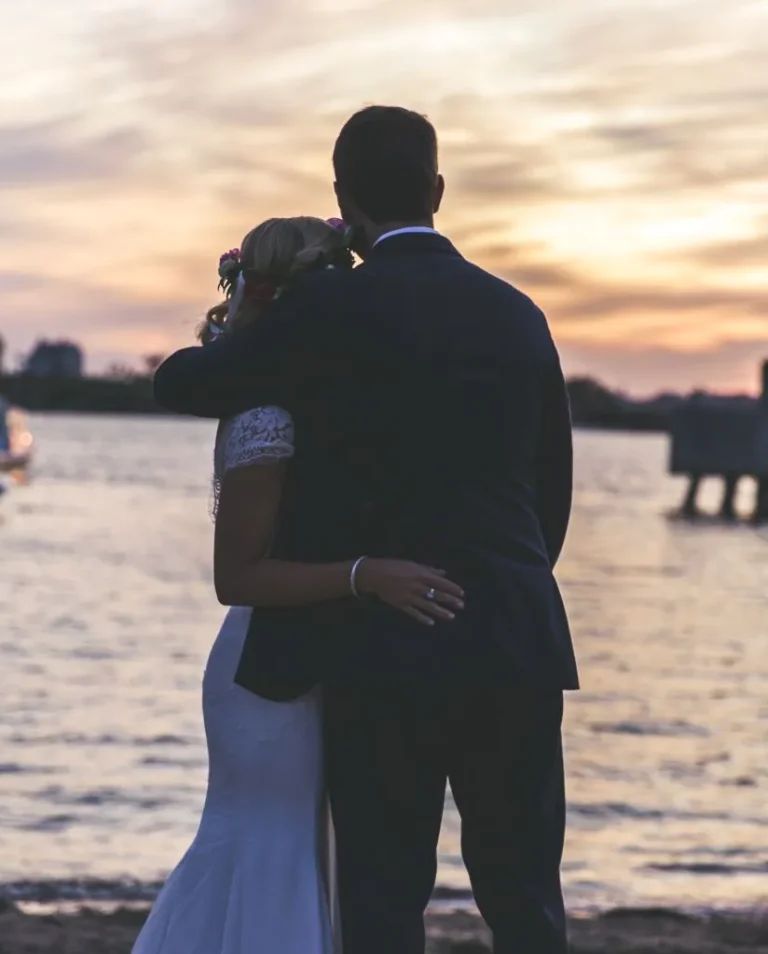 A couple in wedding attire embraces while facing a calm waterfront at sunset, with soft golden and purple hues in the sky and gentle waves in the background.