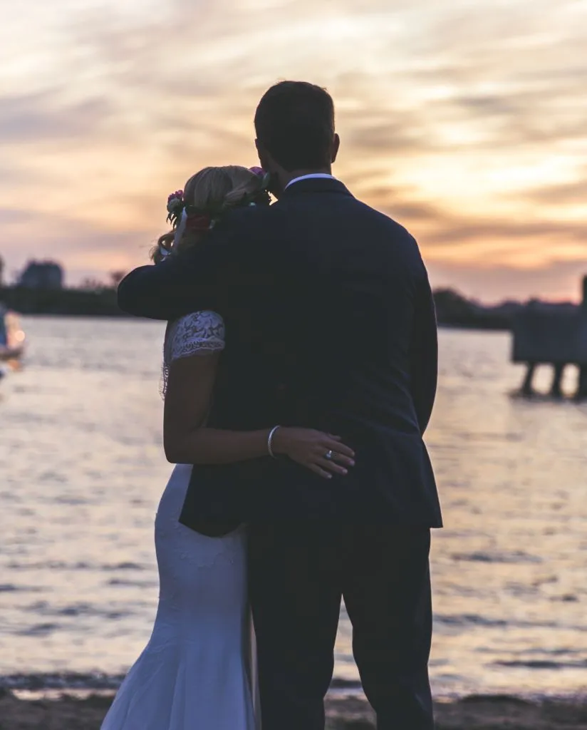 A couple in wedding attire embraces while facing a calm waterfront at sunset, with soft golden and purple hues in the sky and gentle waves in the background.