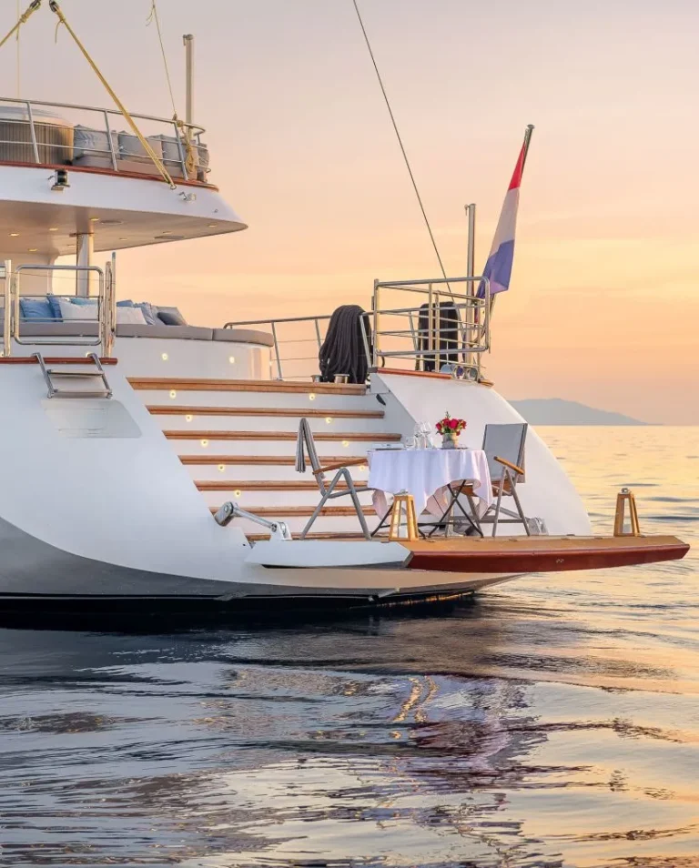 A luxury yacht anchored on calm water at sunset, with a small table and two chairs set for dining on the rear deck. A flag is flying at the stern, and the sky is softly colored in orange and pink hues.