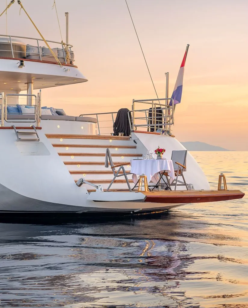 A luxury yacht anchored on calm water at sunset, with a small table and two chairs set for dining on the rear deck. A flag is flying at the stern, and the sky is softly colored in orange and pink hues.