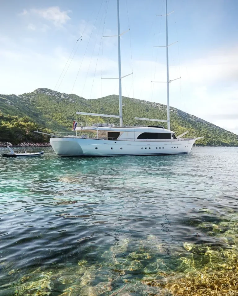 A white yacht with tall masts is anchored in clear, shallow water near a green, hilly coastline under a blue sky. The calm water reveals rocks and pebbles beneath the surface.