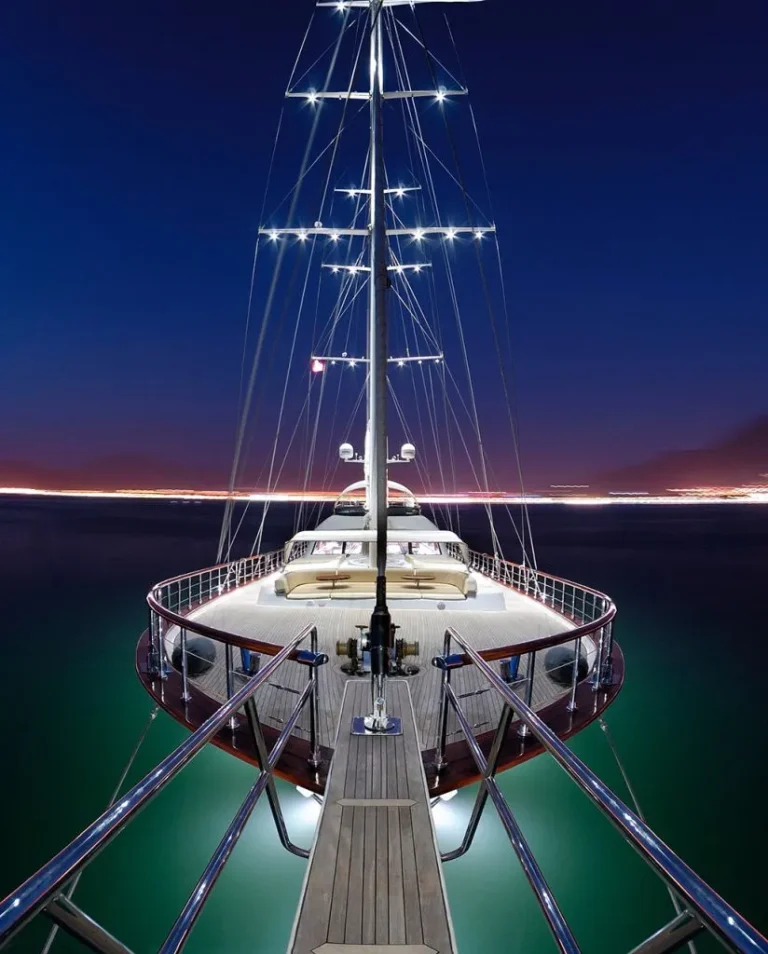 View from the bow of a luxury yacht at night, with deck lights illuminating the area and calm water reflecting the lights. A distant city skyline glows on the horizon under a clear, dark blue sky.