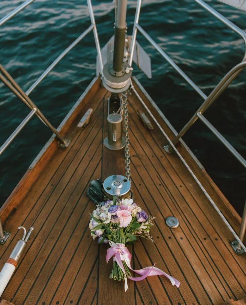 A bouquet of flowers with pink and purple ribbons lies on the wooden deck at the bow of a boat, with deep blue water visible in the background.