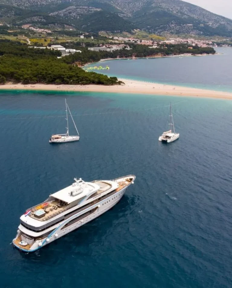 A large yacht and two sailboats are anchored near a narrow, sandy peninsula surrounded by clear blue water, with a forested coastline and hills in the background.