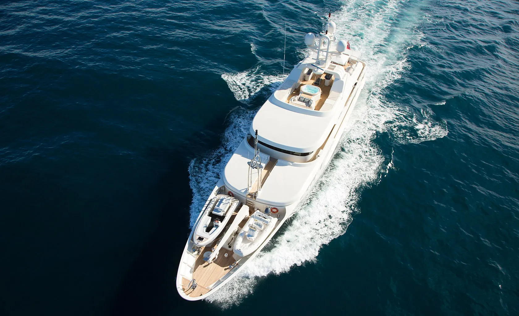 A white luxury yacht cruises through deep blue ocean water, leaving a trail of white waves behind. The yacht has a spacious deck and sleek design, viewed from above.