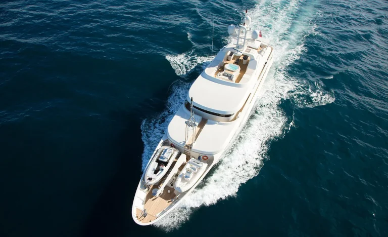 A white luxury yacht travels swiftly through deep blue ocean water, leaving a foamy wake behind it, pictured from above on a sunny day.