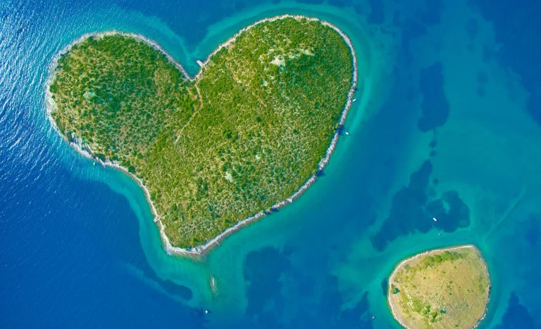 Aerial view of a lush, green island shaped like a heart, surrounded by vibrant blue water, with a smaller, irregularly shaped island nearby.
