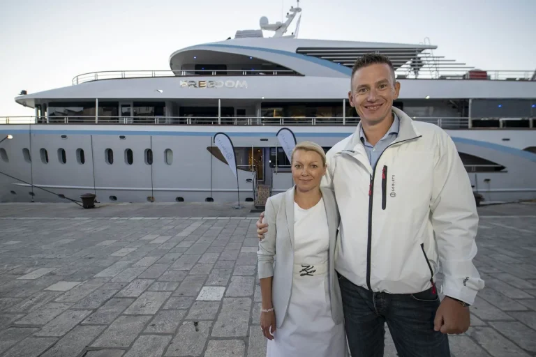 A smiling man and woman stand on a stone dock in front of a large white yacht named FREEDOM during the day. The man has his arm around the woman, and both are wearing light-colored jackets.