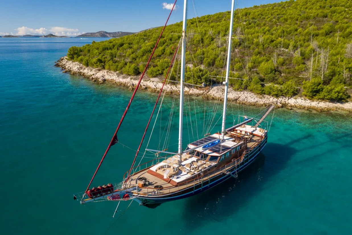 A large sailboat with wooden decks and white lounge chairs is anchored in clear turquoise water near a lush, green, tree-covered coastline under a sunny blue sky.