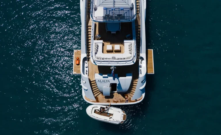 Aerial view of a luxury yacht named ALAYA anchored on blue water, with a smaller boat docked at its stern. Two people are on the smaller boat, and the yacht’s deck has seating areas and sun loungers.
