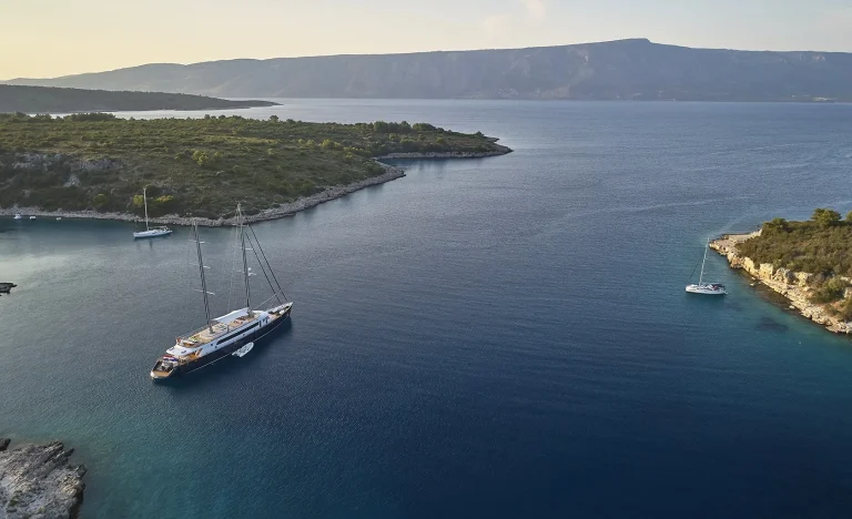 A large sailboat and two smaller boats are anchored in a serene bay surrounded by green hills and calm blue water, with mountains visible in the distance under a clear sky.