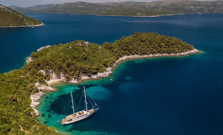 Aerial view of a sailboat anchored near a lush, green island with rocky shores and clear blue water, surrounded by a calm sea and distant hills.