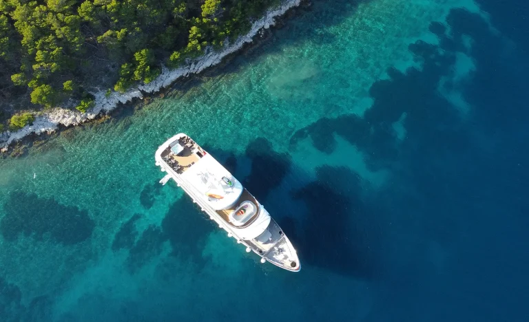 Aerial view of a white yacht anchored near a rocky, tree-lined shore with clear turquoise water gradually deepening to dark blue.
