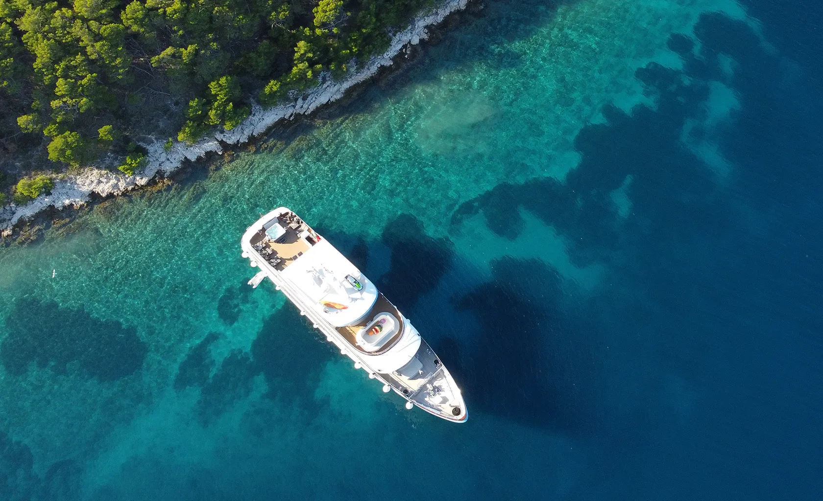 Aerial view of a white yacht anchored near a rocky, tree-lined shore with clear turquoise water gradually deepening to dark blue.