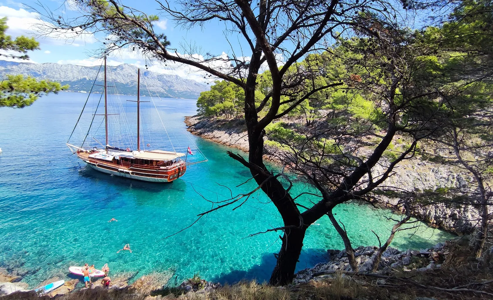 A wooden sailboat is anchored in clear turquoise water near a rocky, tree-lined shore. People swim and paddle near the boat, and the scene is framed by pine trees under a bright blue sky.