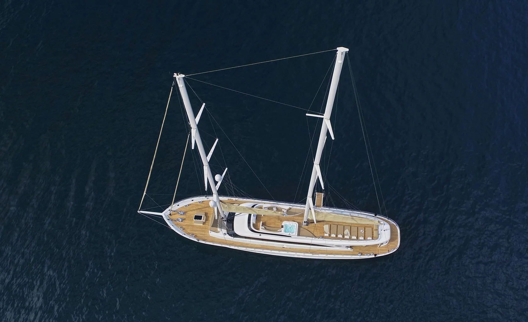 Aerial view of a large sailboat with two masts and a wooden deck floating on calm, dark blue water. The boat features seating areas, a small pool, and open spaces.
