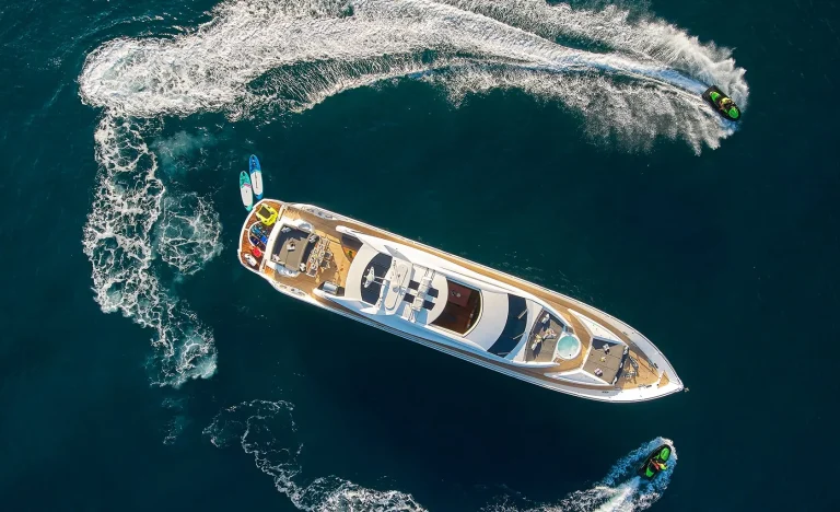 Aerial view of a luxury yacht on deep blue water, surrounded by two jet skis creating circular waves and a small paddleboard floating nearby.