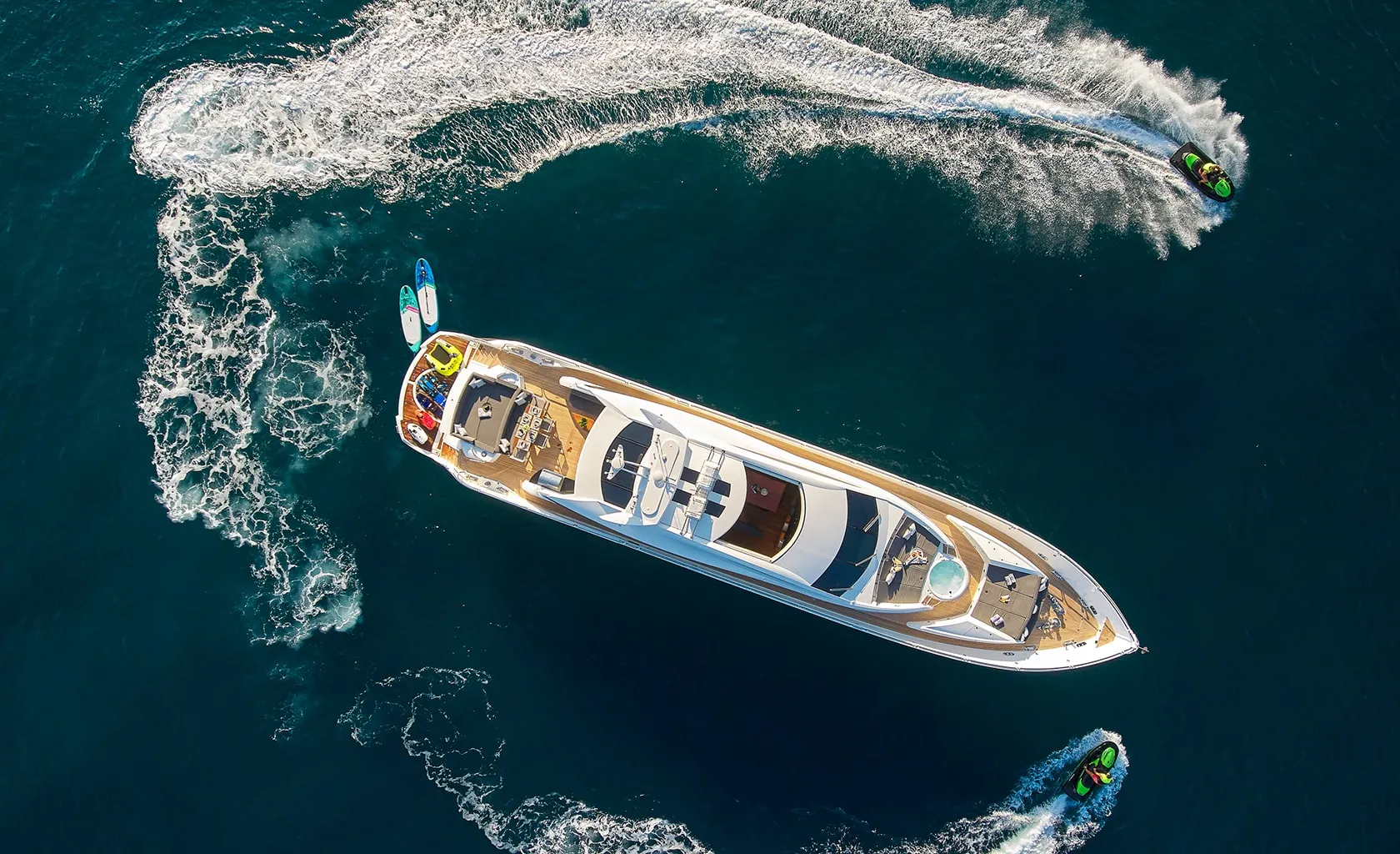 Aerial view of a luxury yacht on deep blue water, surrounded by two jet skis creating circular waves and a small paddleboard floating nearby.