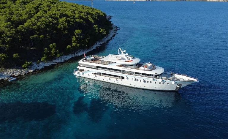 A large white yacht is anchored near a forested coastline with clear blue water, casting a shadow in the sea. The shore curves gently, and the yacht is close to the rocky land covered in dense green trees.