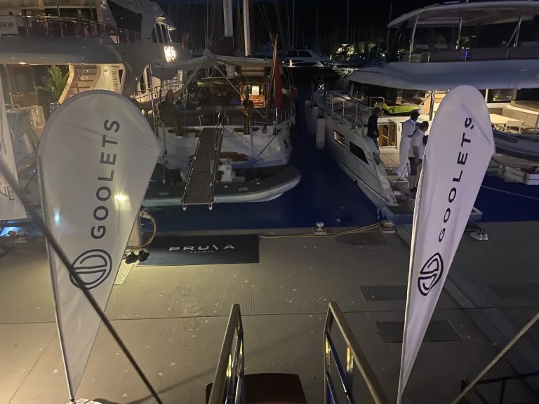 A nighttime marina scene with yachts docked side by side. Two white flags with the GOOLETS logo are visible in the foreground, and the area is softly lit with various lights.