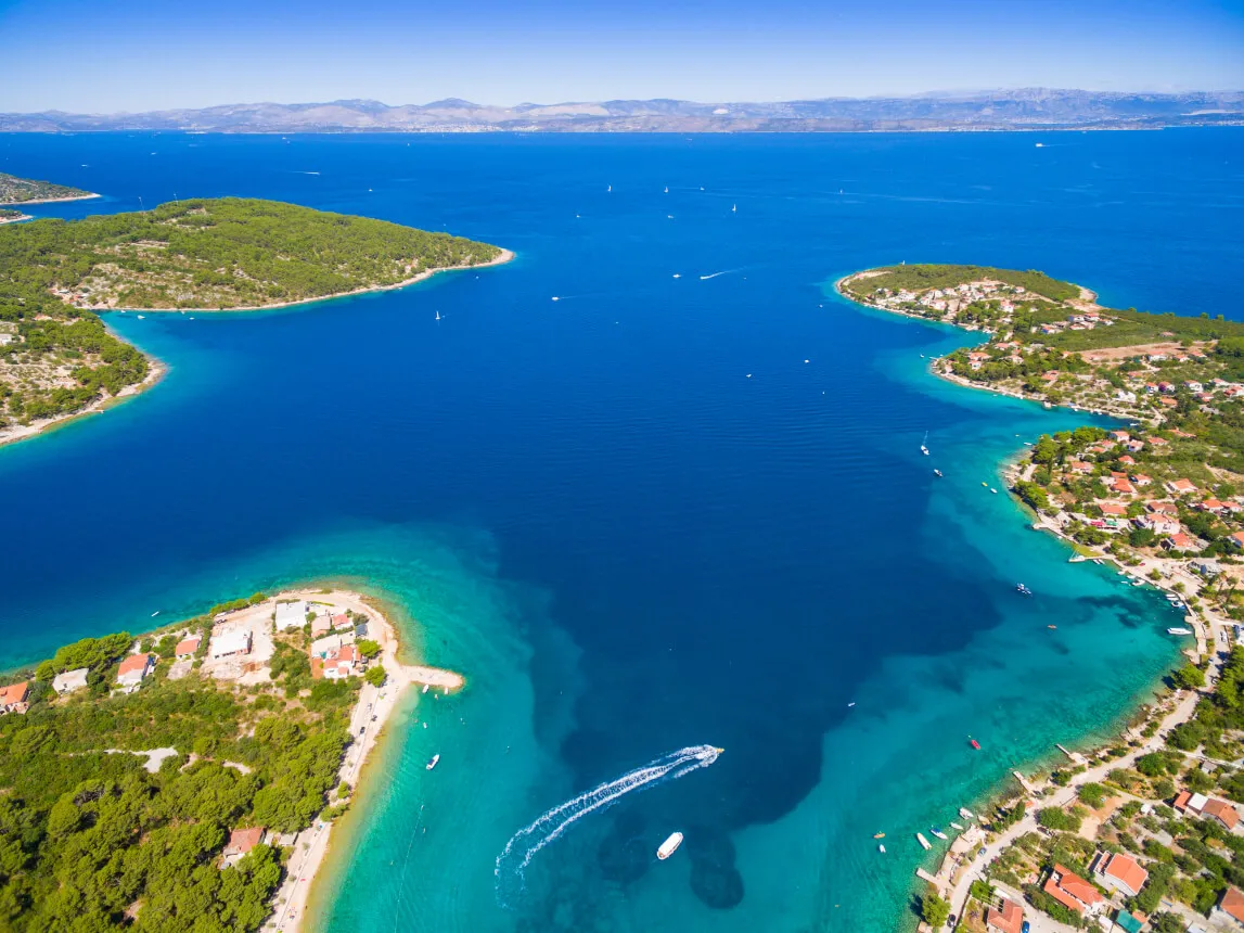 Aerial view of a vibrant blue bay surrounded by green hills and scattered houses, with boats cruising and white wakes visible on the water under a clear sky.