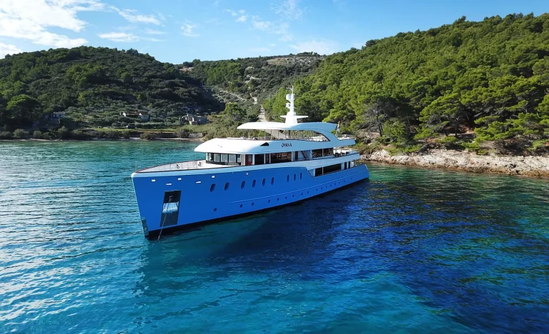 A large, modern yacht with a blue hull is anchored in clear, turquoise water near a rocky, tree-covered coastline under a bright blue sky.