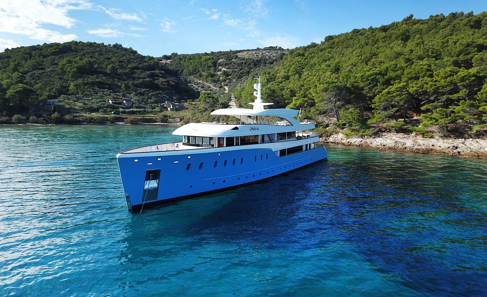 A large, modern yacht with a blue hull is anchored in clear, turquoise water near a rocky, tree-covered coastline under a bright blue sky.
