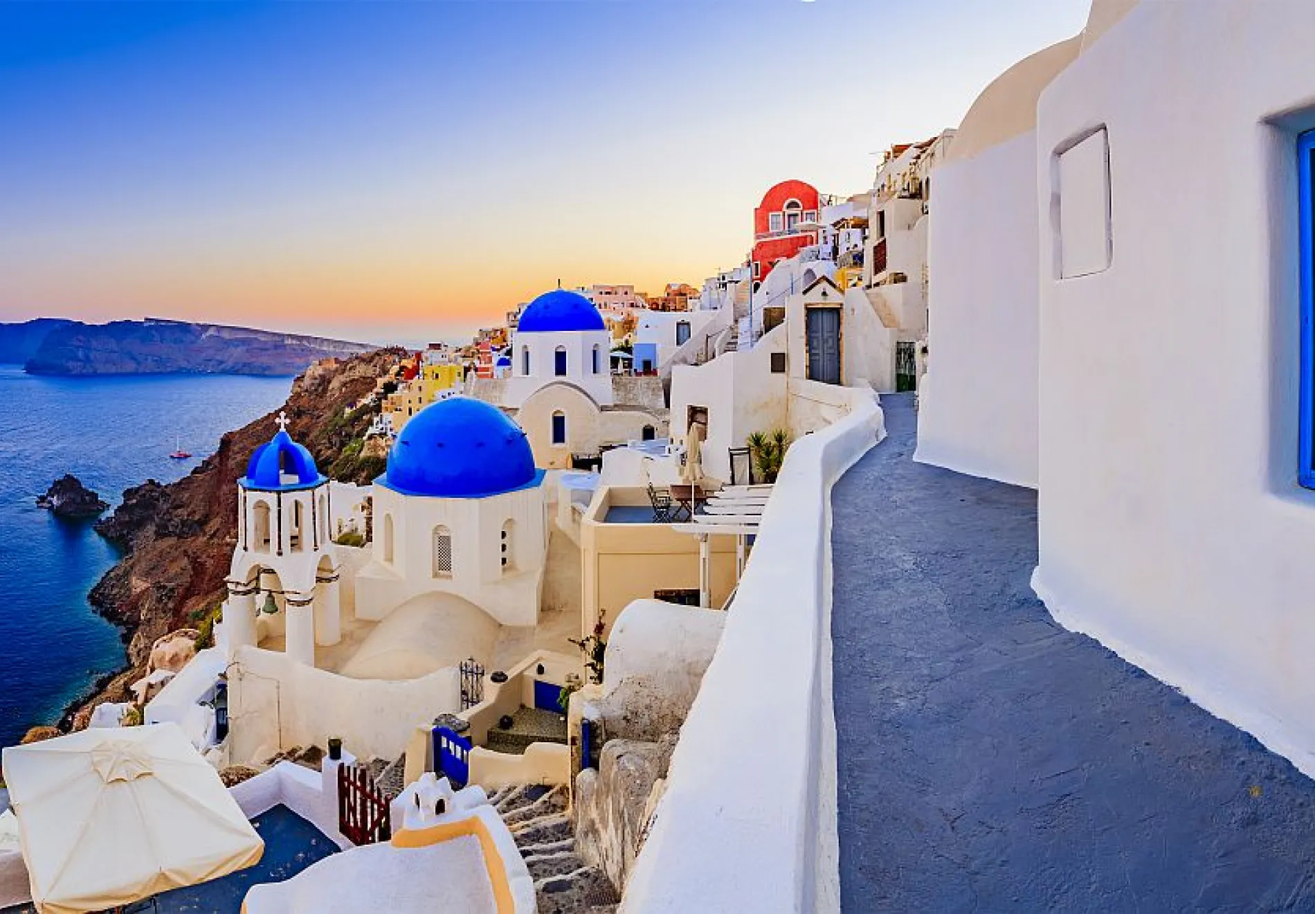 A scenic view of Santorini, Greece at sunset, featuring white-washed buildings with blue domes overlooking the sea and rugged cliffs, with warm light illuminating the architecture.