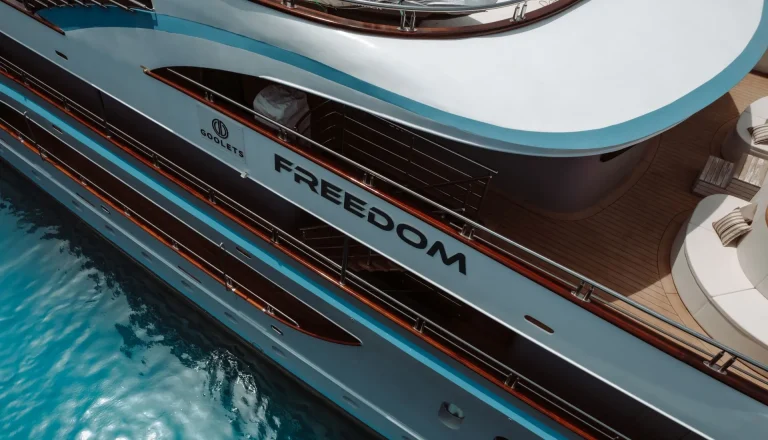 Aerial view of a luxury yacht named FREEDOM docked on clear blue water, displaying sleek white and blue decks with lounging areas and polished railings.