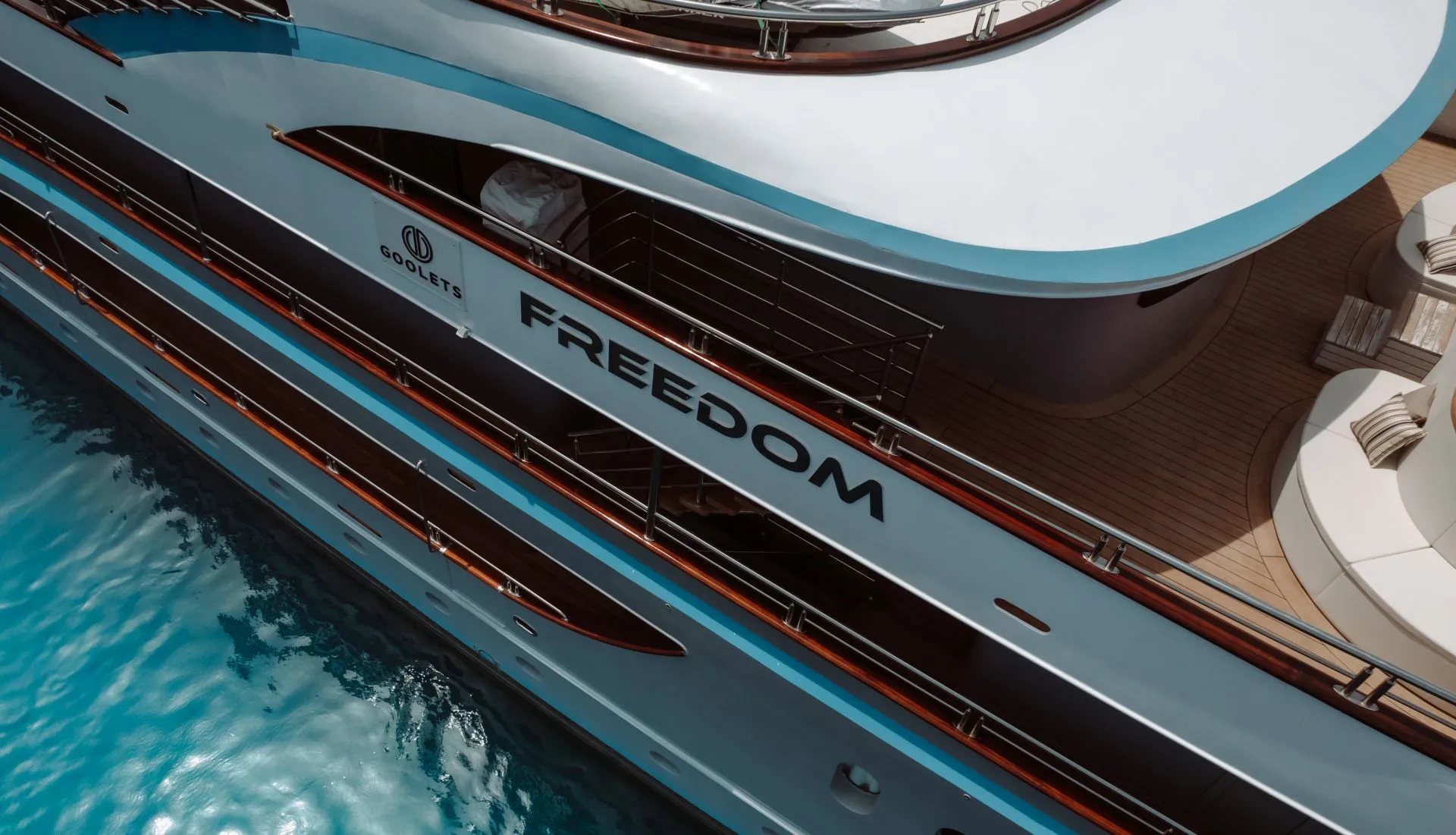 Aerial view of a luxury yacht named FREEDOM docked on clear blue water, displaying sleek white and blue decks with lounging areas and polished railings.