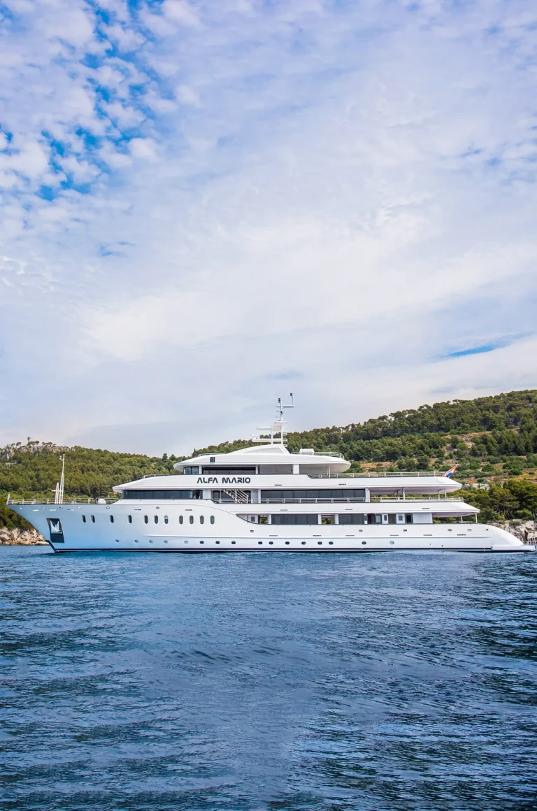 A large white yacht named Alya Mario is anchored near a forested shoreline under a partly cloudy sky, with calm blue water in the foreground.