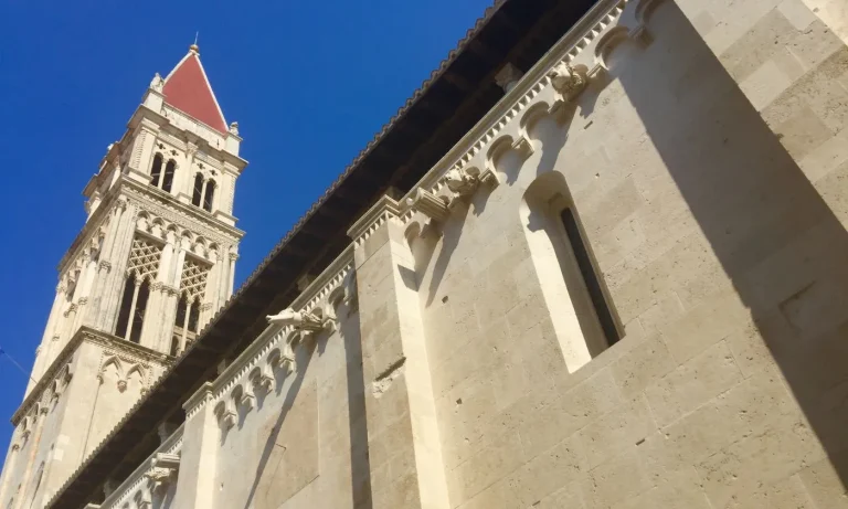 A historic stone church with a tall bell tower featuring a red roof stands against a clear blue sky. The building has arched windows and ornate architectural details along the upper facade.