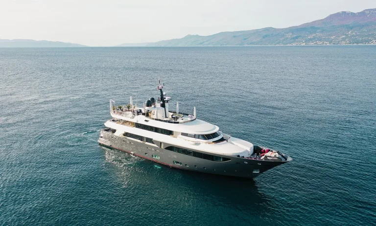 A large luxury yacht sails on calm blue waters near a coastline with hills and mountains in the background under a clear sky.