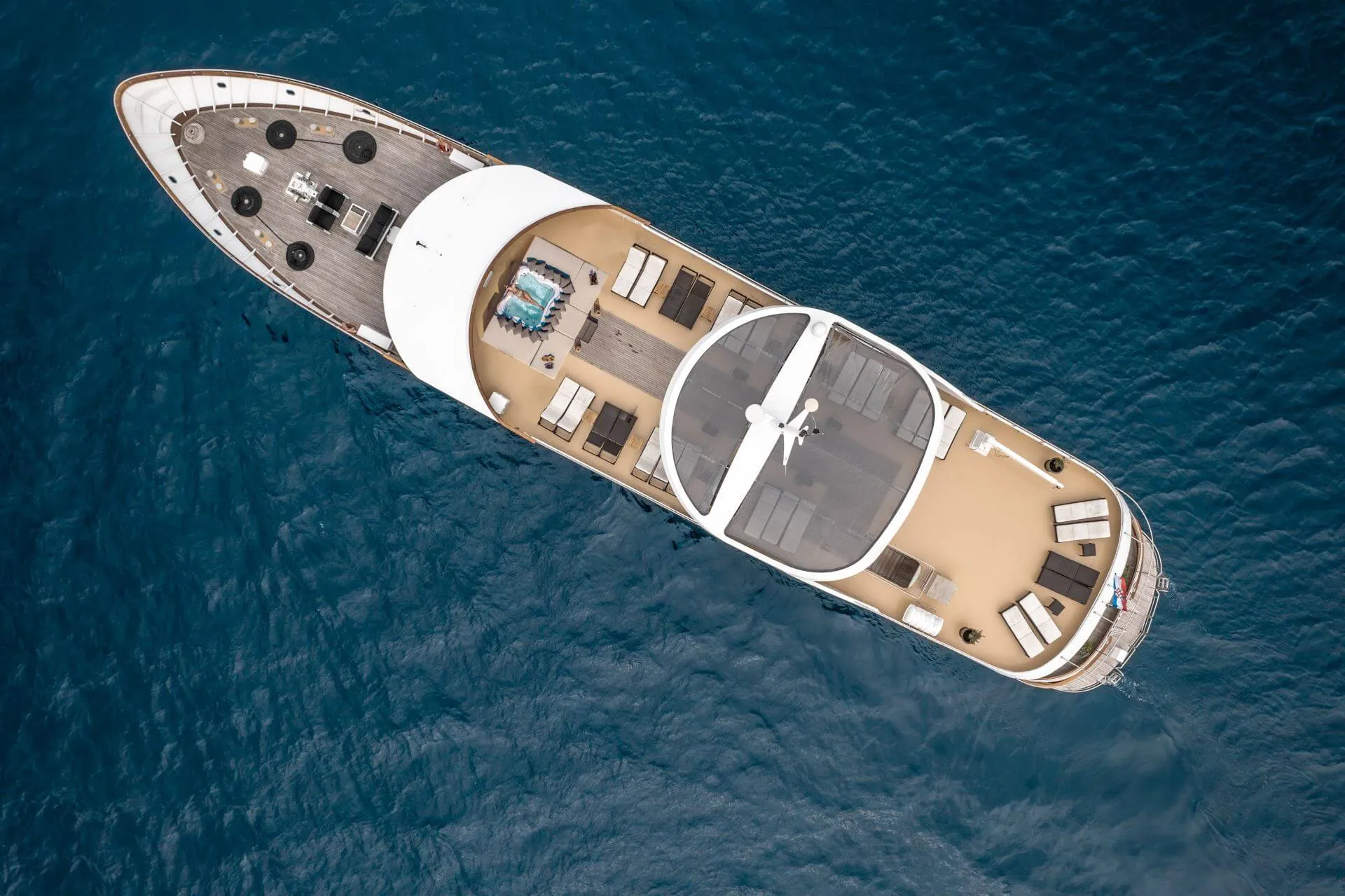 Aerial view of a luxury yacht with sun loungers, seating areas, and a hot tub on the deck, floating on deep blue water.